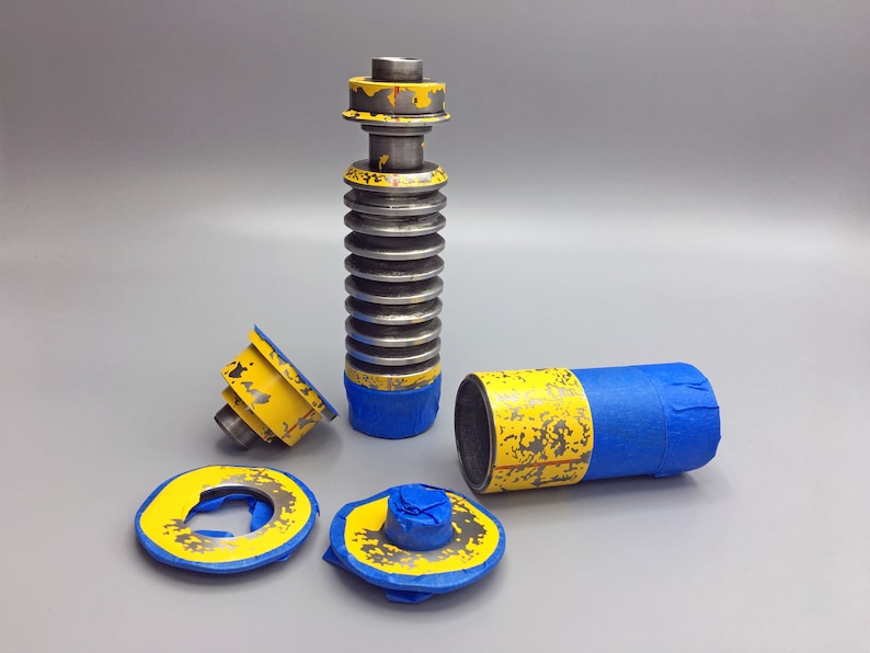 May include: A disassembled lightsaber with a blue and yellow paint scheme. The lightsaber is made of metal and has a textured surface. The lightsaber is in pieces, including the main body, a handle, and a cap.