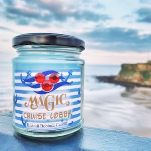 May include: A glass jar candle with a black lid, filled with light blue wax. The label features blue and white stripes, anchors, and the words "MAGIC CRUISE LOBBY." Three red gems are above the text. The candle is on a blue surface with a blurred sea background.