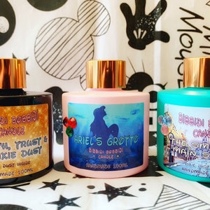 May include: Three glass candle jars with gold lids and labels. The labels feature Disney-themed designs and text. The first jar is black with a Tinkerbell design and the text "Faith, Trust & Pixie Dust". The second jar is pink with an Ariel design and the text "Ariel's Grotto". The third jar is teal with a Main Street, U.S.A. design and the text "The Smell of Main Street".