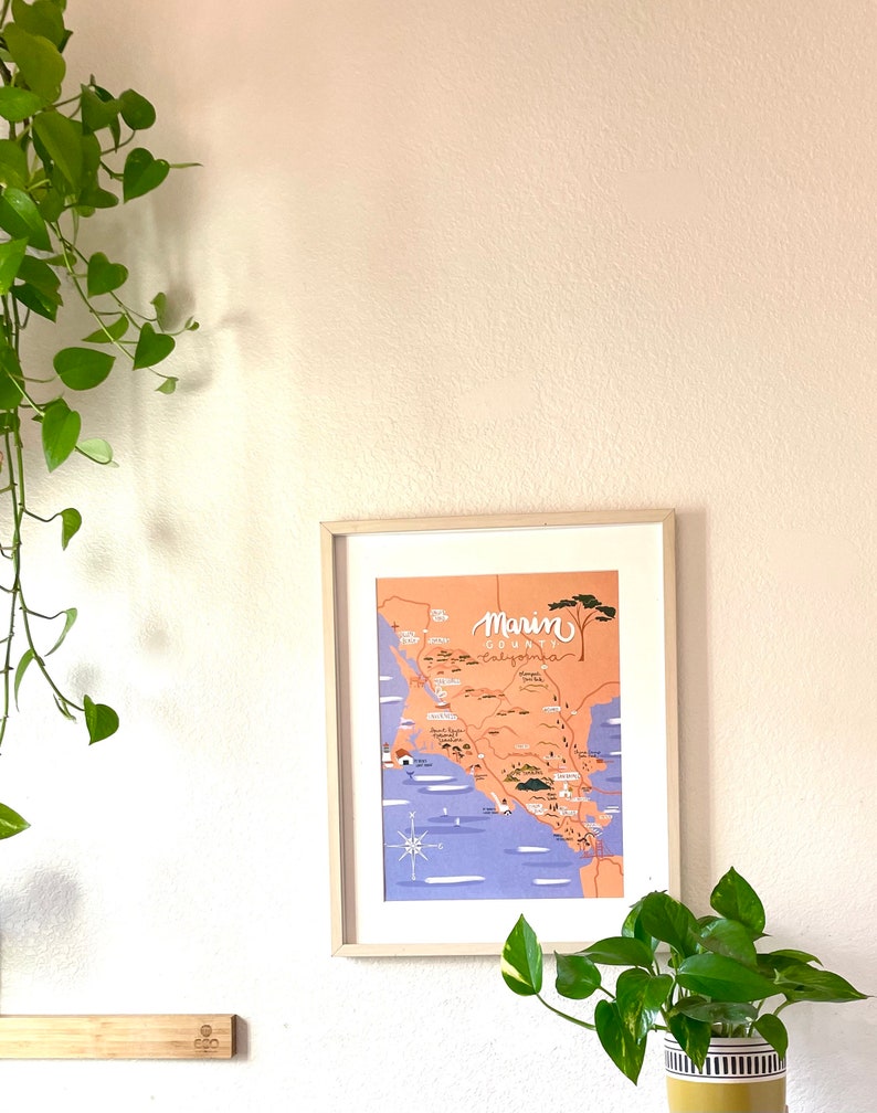 May include: A framed illustration of a map of Marin County, California. The map is in a light peach color with blue water and green trees. The text "Marin County, California" is written in black cursive lettering.