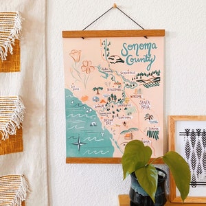 May include: A hand-drawn map of Sonoma County, California, with a pink background and green and blue accents. The map features various towns and cities, including Healdsburg, Geyserville, Windsor, and Santa Rosa. The map is framed with a wooden frame and hanging on a white wall.