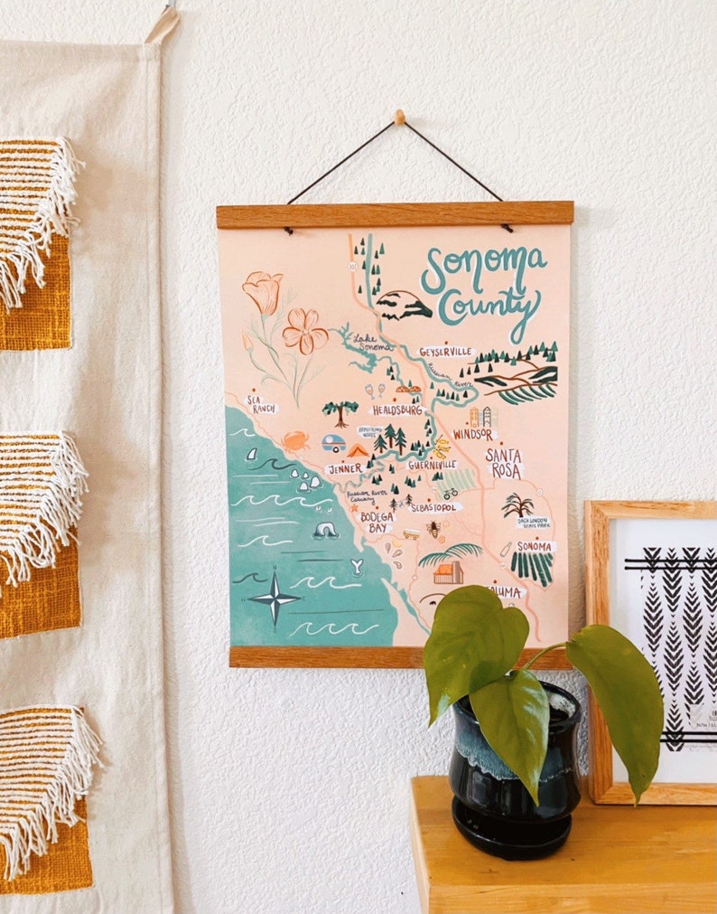 May include: A hand-drawn map of Sonoma County, California, with a pink background and green and blue accents. The map features various towns and cities, including Healdsburg, Geyserville, Windsor, and Santa Rosa. The map is framed with a wooden frame and hanging on a white wall.