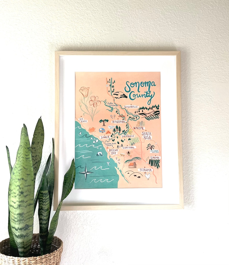 May include: A framed illustrated map of Sonoma County, California. The map is drawn in a whimsical style with hand-drawn lettering and illustrations of landmarks, towns, and natural features. The map is on a peach-colored background with a teal blue border.