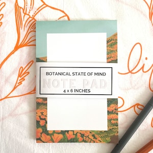 May include: A white notepad with a floral design featuring orange flowers and green leaves. The notepad has a white label with the text "BOTANICAL STATE OF MIND NOTE PAD 4 x 6 INCHES".