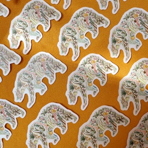 May include: A repeating pattern of white bear-shaped stickers on a yellow background. Each bear is filled with a detailed illustration of various plants, flowers, and leaves in shades of green, brown, yellow, and pink.