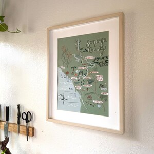 K&ouml;nnte beinhalten: Eine gerahmte Illustration einer Karte von Sonoma County, Kalifornien. Die Karte ist in einem skurrilen Stil mit gr&uuml;nen und braunen Farben gezeichnet. Die Karte enth&auml;lt die Namen verschiedener St&auml;dte und Gemeinden im County, darunter Santa Rosa, Healdsburg und Sonoma.