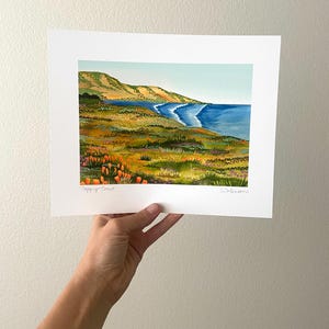Peut inclure: Une peinture &agrave; l'aquarelle d'un paysage c&ocirc;tier avec une mer bleue, une plage de sable et des collines verdoyantes. La peinture pr&eacute;sente un champ de fleurs orange vif au premier plan.