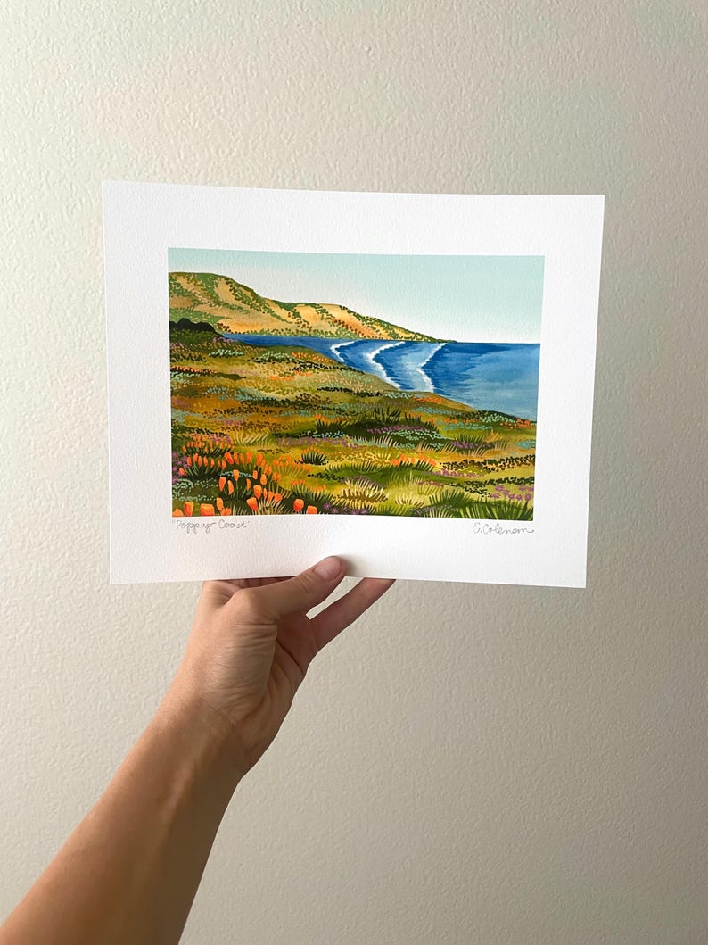 Peut inclure: Une peinture &agrave; l'aquarelle d'un paysage c&ocirc;tier avec une mer bleue, une plage de sable et des collines verdoyantes. La peinture pr&eacute;sente un champ de fleurs orange vif au premier plan.