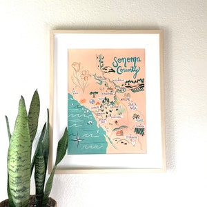 K&ouml;nnte beinhalten: Eine gerahmte Illustration einer Karte von Sonoma County, Kalifornien. Die Karte ist in einem skurrilen Stil mit handgezeichneten Schriftz&uuml;gen und Illustrationen von Sehensw&uuml;rdigkeiten, St&auml;dten und nat&uuml;rlichen Merkmalen gezeichnet. Die Karte befindet sich auf einem pfirsichfarbenen Hintergrund mit einem t&uuml;rkisfarbenen Rand.