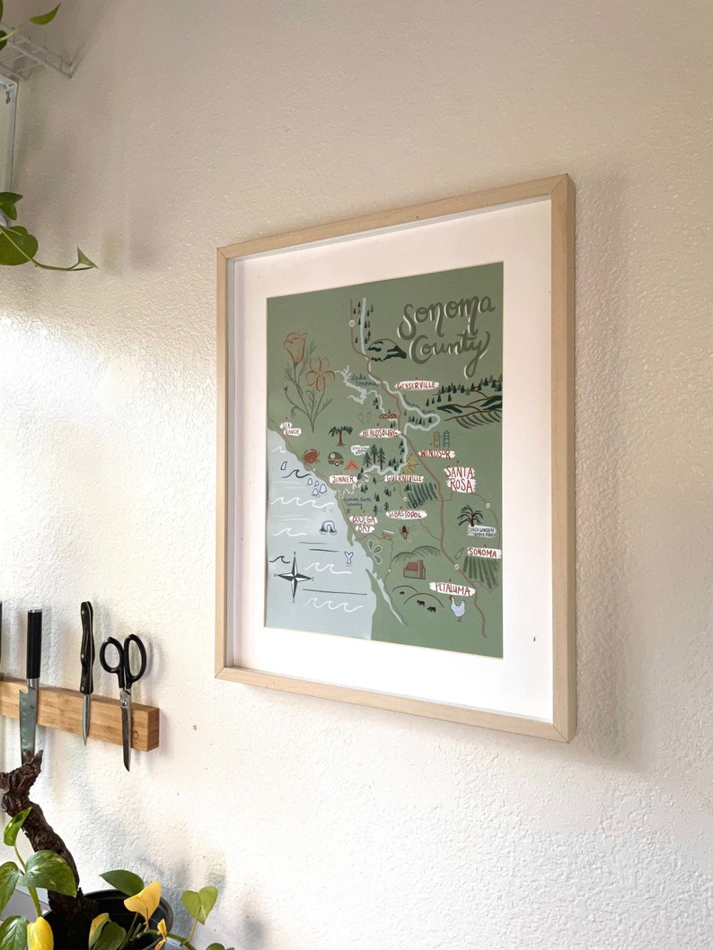 May include: A framed illustration of a map of Sonoma County, California. The map is drawn in a whimsical style with green and blue colors. The map includes the names of various towns and cities in the county, including Santa Rosa, Healdsburg, and Sonoma.
