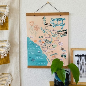 K&ouml;nnte beinhalten: Eine handgezeichnete Karte von Sonoma County, Kalifornien, mit einem pfirsichfarbenen Hintergrund und blauen und gr&uuml;nen Akzenten. Die Karte zeigt die Namen von St&auml;dten und D&ouml;rfern, darunter Santa Rosa, Healdsburg und Bodega Bay. Die Karte ist in einen Holzrahmen gefasst und h&auml;ngt an einer wei&szlig;en Wand.