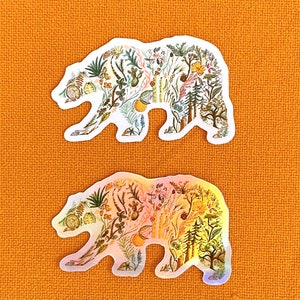 May include: Two bear-shaped stickers with a floral pattern. The top sticker is white with a green, yellow, and brown floral pattern. The bottom sticker is iridescent with a pink, orange, and yellow floral pattern.