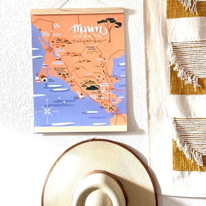 May include: A colorful illustrated map of Marin County, California, with a wooden frame and a string for hanging. The map features various landmarks and towns, including Point Reyes, Sausalito, and Muir Woods. The map is printed on a light blue background with a light orange overlay.