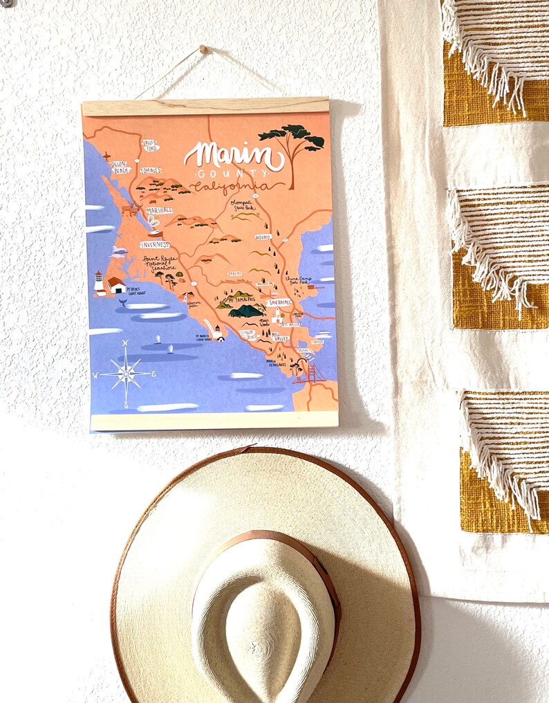 May include: A colorful illustrated map of Marin County, California, with a wooden frame and a string for hanging. The map features various landmarks and towns, including Point Reyes, Sausalito, and Muir Woods. The map is printed on a light blue background with a light orange overlay.