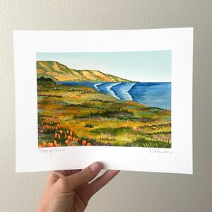 Peut inclure: Une peinture &agrave; l'aquarelle d'un paysage c&ocirc;tier avec une mer bleue, des collines verdoyantes et des fleurs sauvages orange. La peinture est sign&eacute;e "Poppy Coast" et "Coleman".