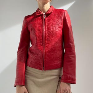 以下が含まれることがあります： 鮮やかな赤色の革製ジャケット。ユニークなスパイラル模様、ジッパー開閉、クラシックな襟が特徴です。ベージュのスカートと組み合わせています。茶色のスタッズ付きハンドバッグが手に持たれています。