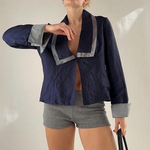 Puede incluir: Blazer azul marino con ribete gris en el cuello y los puños. El blazer está abierto por delante. La modelo lleva shorts grises de canalé. Una bolsa tote negra se sostiene en la mano derecha.