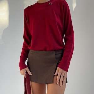May include: A red long-sleeved jumper with a small gold-coloured square detail on the shoulder. It is paired with a brown wrap skirt. A matching red sash is held. The outfit is simple and elegant.