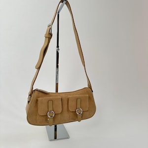 May include: A tan suede shoulder bag with two front pockets, each secured with a flap and a silver-tone ring. The bag has a long, adjustable strap and a zipper closure. The bag is displayed on a silver stand.
