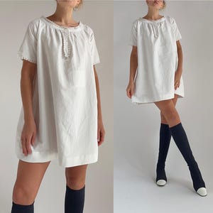 May include: White short-sleeved dress with a round neckline and lace trim. The dress has a loose fit and falls to mid-thigh. The dress features a small embroidered detail on the chest. The model is wearing black knee-high boots.
