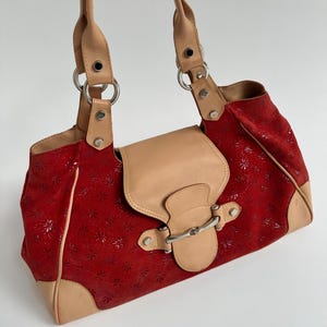 Puede incluir: Bolso de mano rojo con estampado floral y detalles en cuero color canela. El bolso presenta un cierre de solapa con un broche plateado y asas superiores dobles. Las esquinas y la solapa del bolso son de cuero color canela.