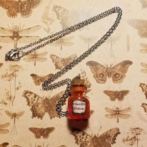 May include: A silver chain necklace with a small glass bottle pendant filled with red glitter. The bottle has a cork stopper and a label that reads "Health Potion".