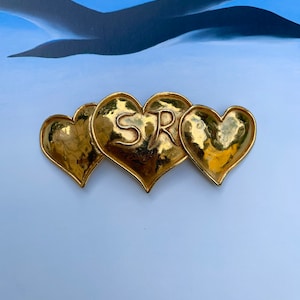 May include: Three gold-tone heart-shaped charms with the letters "SR" embossed on the center charm.