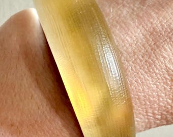 Könnte beinhalten: Ein durchscheinendes, bernsteinfarbenes Armreif-Armband. Das Armband hat eine solide, abgerundete Form und scheint aus einem glatten Material zu bestehen. Das Licht scheint durch das Armband und verleiht ihm ein warmes, leuchtendes Aussehen. Das Armband wird am Handgelenk getragen.