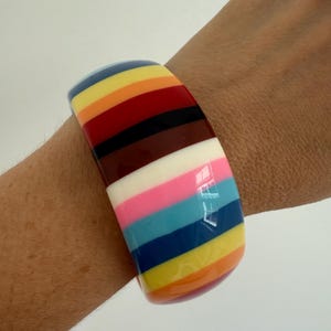 Może przedstawiać: Szeroka, kolorowa bransoletka typu bangle z tęczowym wzorem w paski. Bransoletka posiada paski w kolorze żółtym, pomarańczowym, czerwonym, brązowym, czarnym, białym, różowym, niebieskim i fioletowym. Bransoletka jest noszona na nadgarstku.