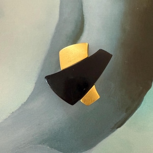 Puede incluir: Un broche moderno con un diseño geométrico. El broche tiene una forma negra angular con dos acentos dorados. La pieza se encuentra sobre un fondo suave y abstracto con tonos de azul y verde.