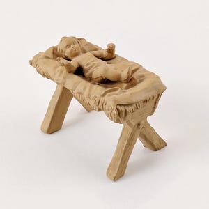 May include: A beige-colored nativity scene figurine depicting a baby lying in a manger. The manger is made of a straw-like material and has wooden legs. The baby is in a relaxed pose.