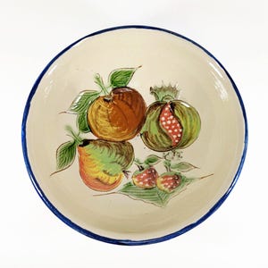 May include: A ceramic bowl with a cream-colored interior and a blue rim. The bowl is decorated with hand-painted fruit, including oranges, pomegranates, and apples with green leaves. The fruit is in shades of brown, green, red, and yellow.