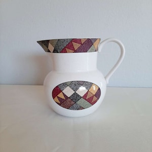 May include: White ceramic pitcher with a geometric pattern in shades of red, green, and brown. The pattern is on the top rim and the center of the pitcher.