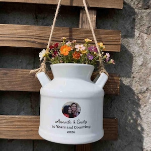 May include: A white ceramic milk can planter hanging by a jute rope, filled with colourful small flowers. The front features a circular photo of two people, with the text Amanda & Emily, 10 Years and Counting, 2026 printed in black ink.
