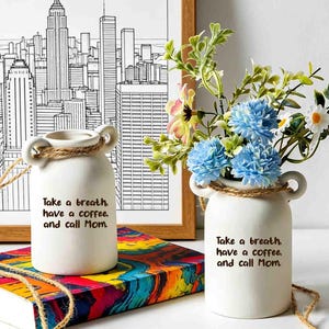 Puede incluir: Dos jarrones de cerámica blanca con dos pequeñas asas y una cuerda rústica. Cada jarrón tiene el texto impreso en negro: Take a breath, have a coffee, and call Mom. Un jarrón está vacío, mientras que el otro contiene un ramo de flores azules y blancas.