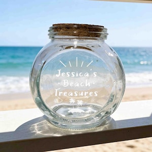 Può includere: Un barattolo di vetro trasparente e rotondo con coperchio in sughero. La parte anteriore del barattolo presenta una scritta bianca che recita "Jessica's Beach Treasures" con un piccolo sole sopra e due palme su un'isola sotto il testo.
