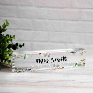 May include: A clear acrylic desk nameplate with the text "Mrs Smith SCHOOL COUNSELOR" in black script. The nameplate is decorated with watercolor-style leaves in shades of green, pink, and peach.