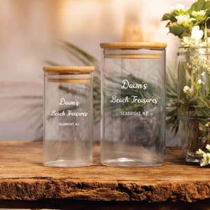 May include: Two clear glass jars with bamboo lids are displayed on a wooden surface. The jars have the text "Dawn's Beach Treasures, SEABRIGHT, NJ" printed on them. The jars are of different sizes, with one taller than the other.