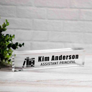 May include: A clear acrylic desk nameplate with the name "Kim Anderson" and the title "Assistant Principal" in black text. A stack of books is to the left of the name. The nameplate is rectangular and sits on a white wooden surface.