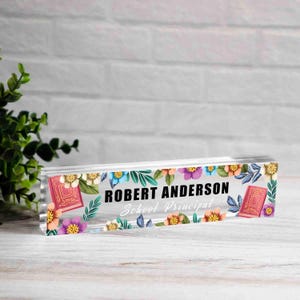 May include: A clear acrylic desk nameplate with floral and leaf accents. The nameplate features the name "ROBERT ANDERSON" and the title "School Principal" in black text. Two red book illustrations are on each side.