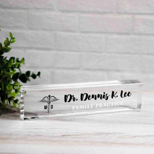 May include: A clear acrylic desk nameplate with the text "Dr. Dennis K. Lee Family Practice" in black script. A medical symbol is also displayed. The nameplate is rectangular and sits on a white wooden surface.
