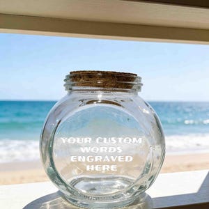Könnte beinhalten: Ein klares, rundes Glasgefäß mit einem Korkstopfen. Auf der Vorderseite des Glases befindet sich der weiß gravierte Text: YOUR CUSTOM WORDS ENGRAVED HERE. Das Gefäß hat eine weite Öffnung und einen flachen Boden zur Aufbewahrung von Objekten.
