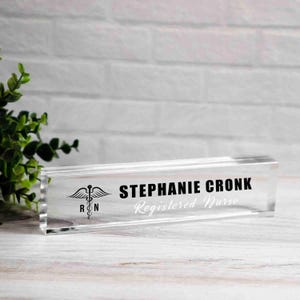 May include: A clear acrylic desk nameplate with the name "STEPHANIE CRONK" in bold black letters and "Registered Nurse" in a script font. A medical symbol is on the left. The nameplate is rectangular and sits on a wooden surface.