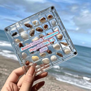 Könnte beinhalten: Ein quadratisches, transparentes Kunststoff-Rasterdisplay mit verschiedenen kleinen Muscheln in einzelnen Fächern. Der Text "Jessica's Beach Treasures Playa Del Rey, CA 2026" ist in blauer und weißer Schrift mittig auf dem Raster aufgedruckt.