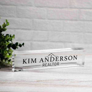 May include: A clear acrylic desk nameplate with the name "KIM ANDERSON" and the title "REALTOR" in black lettering. A small house graphic is above the name. The nameplate is rectangular and sits on a white wooden surface.