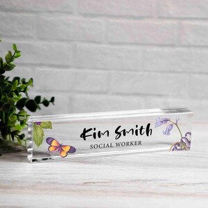 May include: Clear acrylic desk nameplate with the name "Kim Smith" and the title "Social Worker" in black script. A butterfly and floral design adorn the nameplate. The nameplate is rectangular and sits on a white wooden surface.