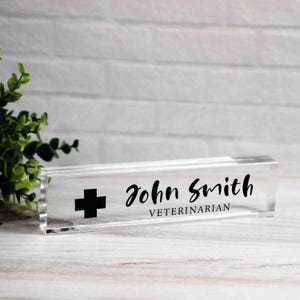 May include: Clear acrylic desk nameplate with the name "John Smith" and the title "Veterinarian" in black script. A black cross symbol is on the left side. The nameplate is rectangular and sits on a white wooden surface.