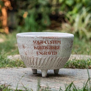 May include: A round, off-white ceramic planter with three short legs and a distressed, textured finish. The front features the brown engraved text: YOUR CUSTOM WORDS HERE ENGRAVED. The planter has a wide, open top and a tapered base.