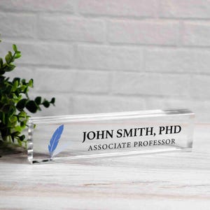 May include: A clear acrylic desk nameplate with the text "JOHN SMITH, PHD ASSOCIATE PROFESSOR" in black. A blue feather graphic is on the left. The nameplate is rectangular and sits on a light wood surface.