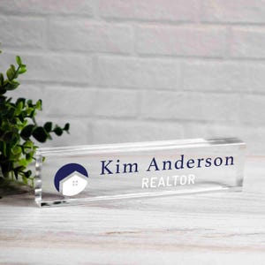 May include: A clear acrylic desk nameplate with the name "Kim Anderson" and the title "REALTOR" in dark blue and white. A blue and white house logo is on the left. The nameplate is rectangular and sits on a white wooden surface.
